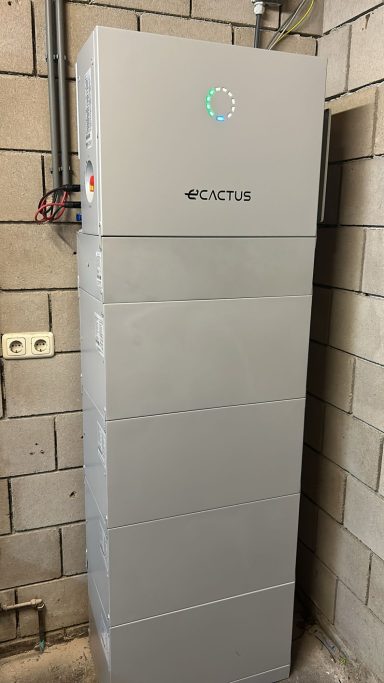 WHES 3-fase batterijpakket 20kWh Grijze energieopslagunit met een groen logo, geplaatst tegen een bakstenen muur.